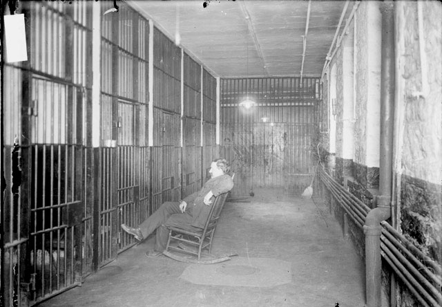 Image of the lockup keeper sitting in a rocking chair outside a row of jail cells in the Harrison Street police station